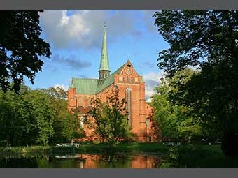 Foto: Gotische Backsteinkirche mit grünem Spitzdach und Turm im Zentrum, hohen, spitzbogigen Fenstern und Verzierungen, umgeben von Bäumen und Grünflächen. Im Vordergrund ein Teich. 