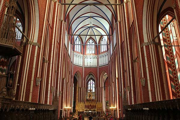 Foto: Innenraum einer gotischen Kathedrale mit roten Ziegelwänden und vertikalen weißen Streifen. Spitzbogiges Gewölbe, unterstützt von filigranen Rippenbögen. Große Fenster im Hintergrund, darunter der geschmückte Altar. 