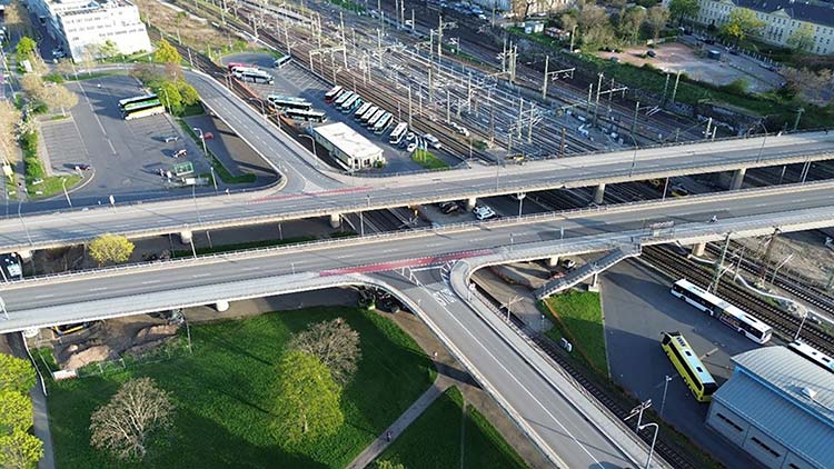 Luftbild, komplexes Verkehrsknotenpunkt-Szenario: Im Zentrum kreuzen sich aufgeständerte mehrspurige Straßenbrücken über einem dichten Bündel parallel verlaufender Eisenbahngleise. 