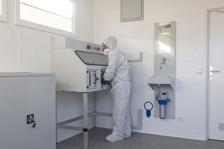 Foto: Eine Person in einem weißen Schutzanzug und Atemschutzmaske steht an einer Apparatur in einem Schadstofflabor. 