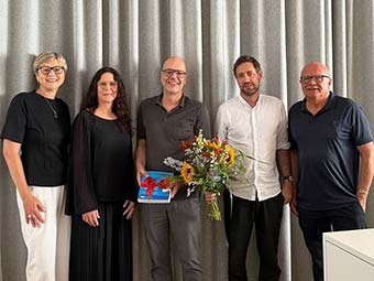 Gruppenfoto: Fünf Personen stehen nebeneinander vor einem grauen Vorhang. Die mittlere Person hält ein blaues Buch mit roter Schleife, die Person rechts daneben einen Blumenstrauß.  