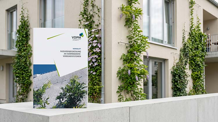 Dien Broschüre mit dem Foto einer begrünten Fassade steht auf einer Betonbrüstung vor einem modernen Wohnhaus mit begrünten Wänden und Balkonen. 