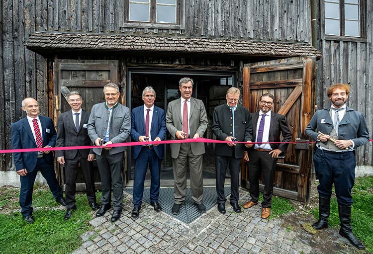 Acht Personen stehen vor einem alten Holzgebäude, vor dem im Rahmen einer Eröffnungsfeier ein rotes Band gespannt ist. Vier der Personen halten eine Schere in der Hand, bereit, das Band durchzuschneiden. 