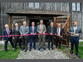 Acht Personen stehen vor einem alten Holzgebäude, vor dem im Rahmen einer Eröffnungsfeier ein rotes Band gespannt ist. Vier der Personen halten eine Schere in der Hand, bereit, das Band durchzuschneiden. 