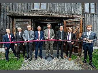 Acht Personen stehen vor einem alten Holzgebäude, vor dem im Rahmen einer Eröffnungsfeier ein rotes Band gespannt ist. Vier der Personen halten eine Schere in der Hand, bereit, das Band durchzuschneiden. 