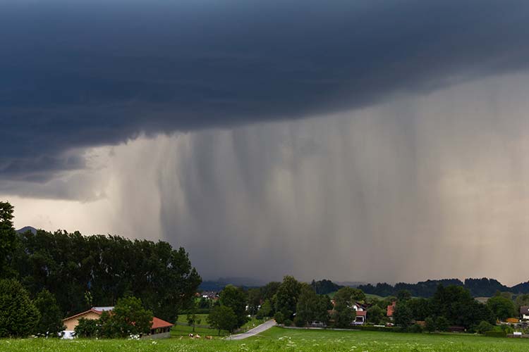 Starker Regenschauer unter dunklen Gewitterwolken über einer grünen Landschaft mit verstreuten Häusern und Bäumen. 