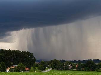 Starker Regenschauer unter dunklen Gewitterwolken über einer grünen Landschaft mit verstreuten Häusern und Bäumen. 