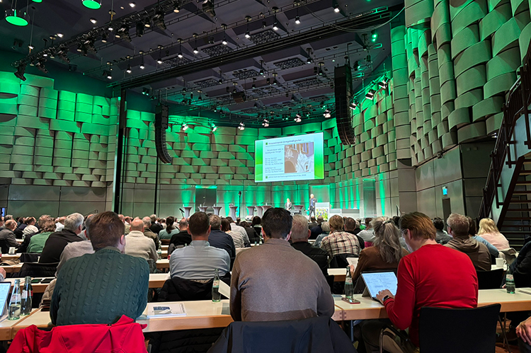Das Bild zeigt einen großen Konferenzraum mit vielen Teilnehmern, die an Tischen sitzen. Im Hintergrund ist eine große Leinwand zu sehen, auf der eine Präsentation läuft. Die Wände sind mit akustischen Paneelen verkleidet und in grünem Licht beleuchtet. AIBau GmbH