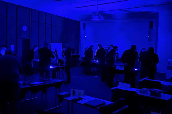 Ein abgedunkelter Seminarraum mit mehreren Personen, die in kleinen Gruppen an Arbeitstischen stehen. Die Tische sind mit Dokumenten und Geräten bestückt. Der Raum ist in blauem Licht beleuchtet. 
