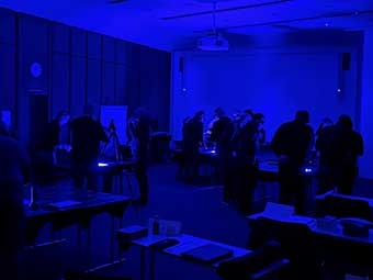 Ein abgedunkelter Seminarraum mit mehreren Personen, die in kleinen Gruppen an Arbeitstischen stehen. Die Tische sind mit Dokumenten und Geräten bestückt. Der Raum ist in blauem Licht beleuchtet. 