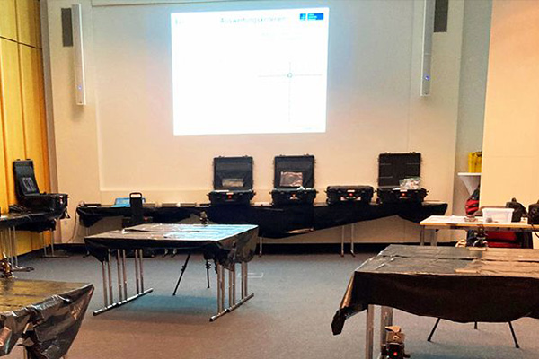Foto:  Seminarraum mit Messtechnik. Im Vordergrund mehrere rechteckige Tische mit schwarzer Abdeckung. Im Hintergrund vor einer Projektionsfläche steht eine Reihe geöffneter schwarzer Transportkoffer mit Geräten. 
