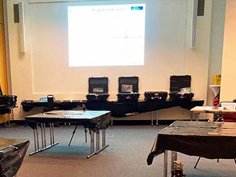 Foto:  Seminarraum mit Messtechnik. Im Vordergrund mehrere rechteckige Tische mit schwarzer Abdeckung. Im Hintergrund vor einer Projektionsfläche steht eine Reihe geöffneter schwarzer Transportkoffer mit Geräten. 