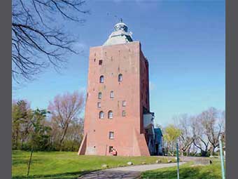 Foto: Leuchtturm mit quadratischem Grundriss und flacher Spitze, auf der sich eine Laterne befindet. Das Gebäude hat mehrere Fenster unterschiedlicher Größe und steht in einer Grünanlage mit Bäumen.  