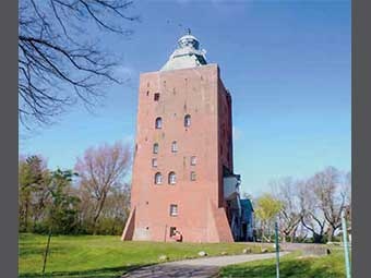 Foto: Leuchtturm mit quadratischem Grundriss und flacher Spitze, auf der sich eine Laterne befindet. Das Gebäude hat mehrere Fenster unterschiedlicher Größe und steht in einer Grünanlage mit Bäumen.  