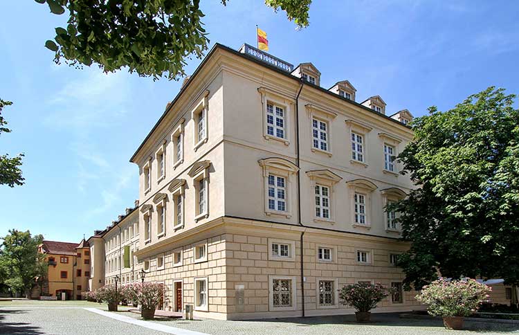 Foto: Mehrstöckiges, hellbeiges Gebäude im klassischen Architekturstil mit symmetrischer Fensteranordnung auf allen Etagen. 