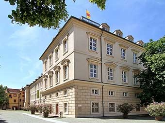 Foto: Mehrstöckiges, hellbeiges Gebäude im klassischen Architekturstil mit symmetrischer Fensteranordnung auf allen Etagen. 