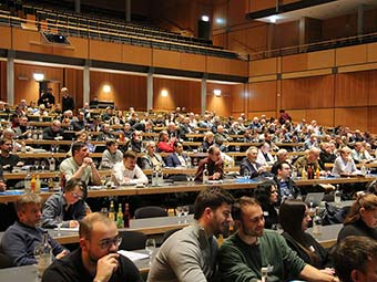 Foto, Innenraum, Großveranstaltung: Mehrere hundert Personen sitzen in einem mehrreihigen Hörsaal an langen Tischen und hören einer Veranstaltung zu, einige  machen Notizen oder arbeiten an Laptops; auf den Tischen stehen Wasserflaschen und Getränke, der Raum ist holzvertäfelt und gut beleuchtet. 