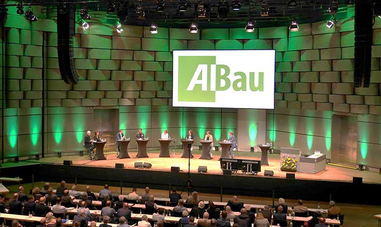 Großer Veranstaltungssaal mit 7 Pulten, an denen Redner stehen. Blick aus den Publikumsreihen zur Bühne. 